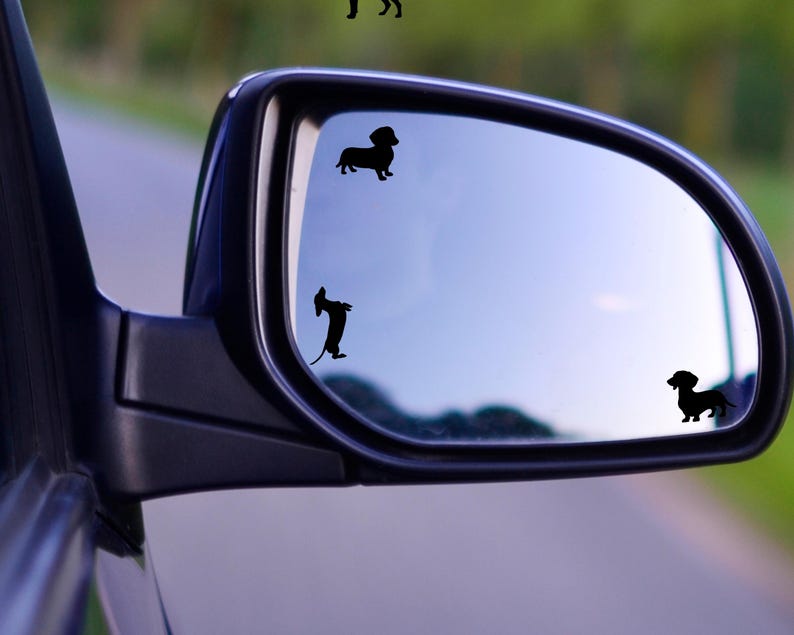 May include: A rearview mirror with three black dachshund silhouette decals. The decals are on the mirror's surface, reflecting the sky and road.