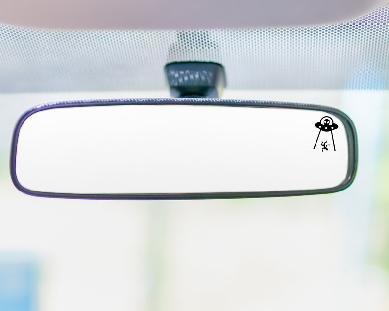 May include: A black rearview mirror with a small black sticker of a UFO abducting a person. The sticker is in the upper right corner of the mirror.