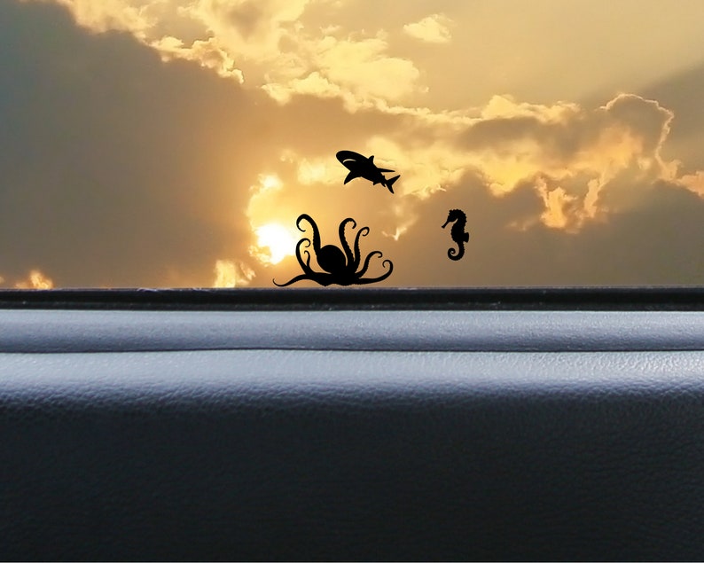 May include: Silhouette of an octopus, a shark, and a seahorse against a sunset sky. The silhouettes are black and are placed on a car window.
