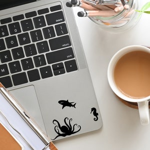 May include: A silver laptop computer with a black keyboard and a black octopus, shark, and seahorse sticker on the lid. The laptop is on a white surface with a cup of coffee and a pencil holder.