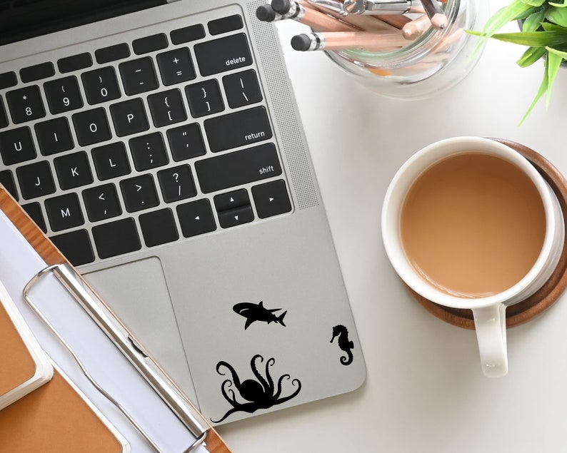 May include: A silver laptop computer with a black keyboard and a black octopus, shark, and seahorse sticker on the lid. The laptop is on a white surface with a cup of coffee and a pencil holder.