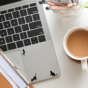 Puede incluir: Un ordenador port&aacute;til plateado con un teclado negro y un trackpad negro. Hay tres pegatinas de gato negro en el trackpad. Una taza de caf&eacute; est&aacute; a la derecha de la imagen.