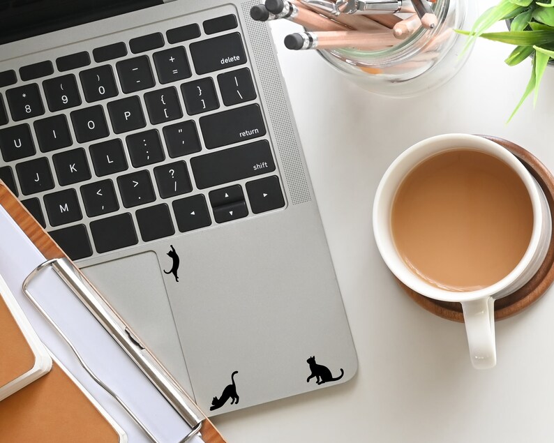 Puede incluir: Un ordenador port&aacute;til plateado con un teclado negro y un trackpad negro. Hay tres pegatinas de gato negro en el trackpad. Una taza de caf&eacute; est&aacute; a la derecha de la imagen.