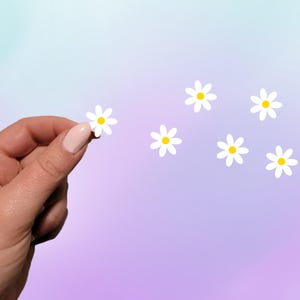 Puede incluir: Una mano sosteniendo una margarita blanca con un centro amarillo, con varias otras margaritas dispersas sobre un fondo degradado suave púrpura y azul. Las margaritas tienen un diseño simple y limpio.