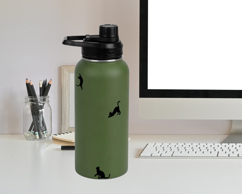 Puede incluir: Una botella de agua reutilizable verde con tapa y asa negras. La botella tiene tres siluetas de gatos negros en ella.