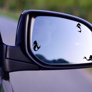 May include: A rearview mirror with three black mermaid silhouettes on the reflective surface. The mirror is attached to a car.