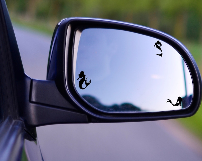 May include: A rearview mirror with three black mermaid silhouettes on the reflective surface. The mirror is attached to a car.