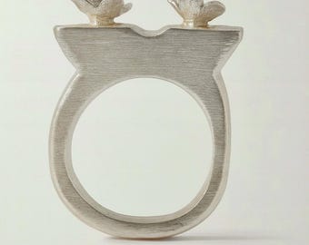 May include: A silver-toned ring featuring a floral design. The ring has a wide band with a large circular opening. Two small flower-shaped embellishments sit atop the ring's upper portion. The ring is made of a brushed metal.