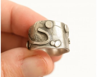 Puede incluir: Un anillo ancho de color plateado con un diseño abstracto. El anillo presenta áreas elevadas y texturizadas, así como adornos circulares. También hay una forma curva y estilizada. El anillo está sostenido por los dedos de una persona.