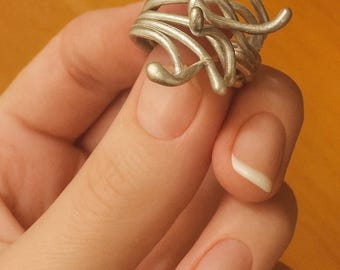 May include: A silver-colored ring with an abstract design. The ring features multiple curved and looped metal elements, creating a unique and artistic appearance. The ring is held in a hand with manicured nails, showcasing the jewelry's details.
