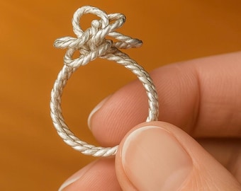 May include: A silver-colored ring with a knot design. The ring's band and knot are made of a twisted, rope-like material. The ring is held by a person's fingers against an orange background.