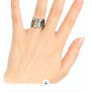 May include: A silver-toned ring with a textured design and circular accents is displayed on a hand with manicured nails. The ring appears to be made of metal and has a unique, artistic style.
