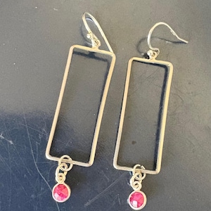 May include: A pair of silver-toned rectangular drop earrings. Each earring features a rectangular frame with a small, round, red gemstone dangling from the bottom. The earrings have a simple, elegant design.