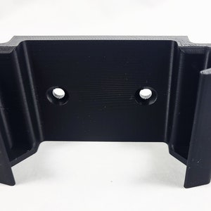 May include: Black plastic wall mount with two holes for screws. The mount has a U-shaped opening for holding an object.