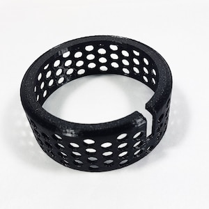 May include: A black, 3D-printed ring with a series of evenly spaced, circular holes. The ring has a gap on one side. The object is on a white surface.