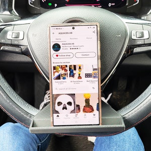 May include: A smartphone displaying the Etsy shop AQUA3DLAB, featuring 3D printed items, including a skull, a cactus, and a reef aquarium. The phone is resting on a grey phone holder attached to a car's steering wheel.