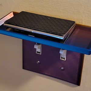 Könnte beinhalten: Ein kleines, blaues Glasregal, das an einer Lichtschalterplatte befestigt ist. Eine schwarze Brieftasche liegt auf dem Regal.