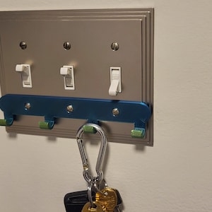 Könnte beinhalten: Ein blauer und grüner Schlüsselhalter mit Haken, der an einer Lichtschalterplatte befestigt ist. Ein Karabiner mit Schlüsseln hängt an einem der Haken.
