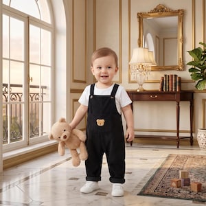 May include: A toddler wearing a white short-sleeved t-shirt and black corduroy overalls with a small embroidered bear on the chest pocket. The child is holding a light brown plush teddy bear and wearing white sneakers on a marble floor.