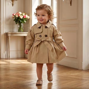 May include: A beige trench coat featuring a double-breasted front with dark brown buttons, a wide collar, puffed long sleeves with cuffs, and a matching fabric tie belt at the waist. The coat has a flared skirt silhouette and is made of smooth fabric.