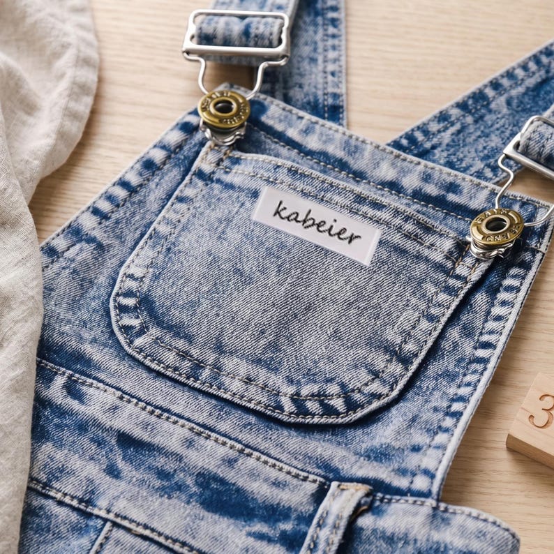 May include: Close-up of a denim overall with a light blue acid wash. The overall features adjustable straps with metal hardware and a front pocket with the brand name "kabeier" on a white label. The fabric has a textured appearance.