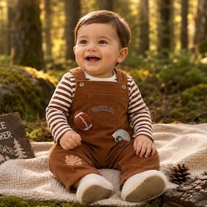May include: A smiling baby wearing a brown corduroy overall with football and helmet patches, paired with a striped long-sleeve shirt. The outfit includes white, fluffy-lined booties. The baby is sitting on a blanket in a forest setting.