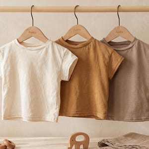 Puede incluir: Tres camisetas de manga corta en crema, mostaza y topo cuelgan de una barra de madera. Cada camisa está en una percha de madera con el nombre "Aosta" impreso. Las camisetas se exhiben sobre un fondo neutro, con juguetes de madera y un paño doblado en primer plano.