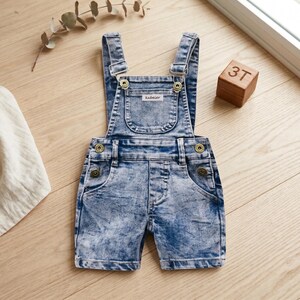May include: Light blue denim overalls with a stonewashed effect. The overalls have adjustable straps, a bib pocket with the brand name "kabeier", and brass-colored buttons. A wooden block with the number "3T" is also in the image.