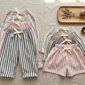 Puede incluir: Una colección de pantalones y shorts a rayas con cinturas elásticas y cordones de ajuste en color crema. Las prendas se presentan en varias combinaciones de colores, incluyendo rayas verticales grises, rosas y beige sobre un fondo claro.