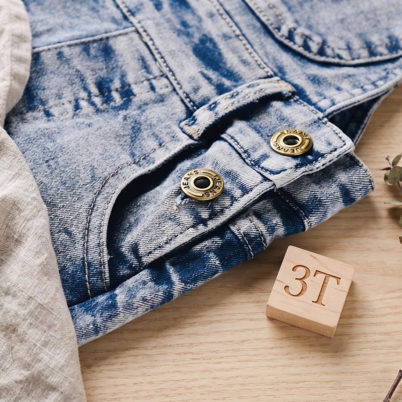 May include: Close-up of a light blue denim garment with brass-colored buttons that read "JEANS". A wooden block with the number "3T" is also visible. The denim has a faded, textured appearance.