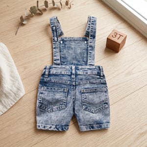 May include: Light blue denim overalls with a stonewashed effect. The shorts-style overalls have adjustable straps and pockets. A wooden block with the number "3T" is visible in the background.