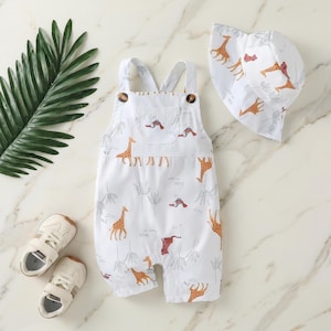 May include: A white sleeveless romper with a giraffe and tree print, featuring two brown buttons on the straps, a matching white bucket hat with a giraffe print, and a pair of white hook-and-loop closure sneakers for a complete outfit.