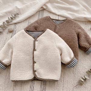 May include: Two long-sleeved jackets made of textured fleece material. One jacket is cream-coloured and the other is brown. Both feature a V-neckline, a front button closure, and a black and white gingham check pattern lining visible at the cuffs.