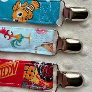 May include: Four fabric pacifier clips with silver metal clasps. The top strap is yellow with cartoon characters. The second is turquoise with an orange fish. The third is light blue with a rooster. The bottom strap is red with race car graphics.