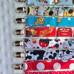 May include: A collection of eight fabric pacifier clips, each featuring a silver metal clasp. The straps display various patterns including cartoon characters, race cars, ice cream, Mickey Mouse silhouettes, and red polka dots of different sizes.