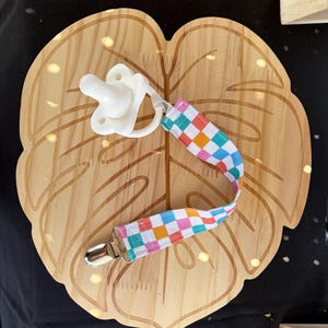 May include: A white pacifier attached to a fabric strap featuring a multi-colored checkered pattern in pink, orange, blue, and green. The strap includes a silver metal clip at one end for attachment, providing a secure way to hold the pacifier.