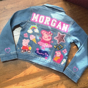 May include: A light blue denim jacket with the name "MORGAN" in pink letters across the back. The jacket is decorated with Peppa Pig patches, a rainbow, a star, musical notes, and hearts.