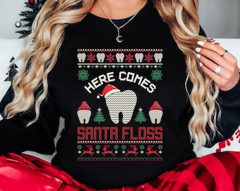 May include: Black crew neck sweatshirt with a festive Christmas design. The design includes a tooth wearing a Santa hat, the text "HERE COMES SANTA FLOSS", teeth, snowflakes, Christmas trees, and reindeer. The sweatshirt is a holiday-themed item.
