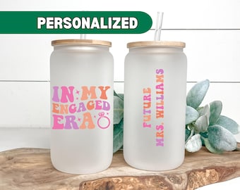 May include: Two frosted glass tumblers with lids and straws. One tumbler says "In My Engaged Era" with a pink, orange, and purple font and a diamond ring graphic. The other tumbler says "Future Mrs. Williams" with a pink, orange, and purple font.