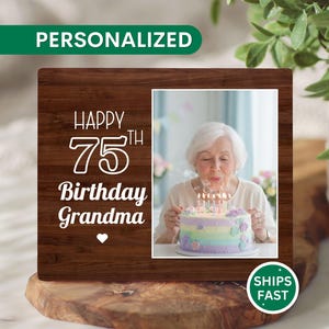 May include: A personalized wooden picture frame with the text "HAPPY 75th Birthday Grandma" in white. The frame holds a photo of a woman blowing out candles on a birthday cake. The frame is on a wooden surface.