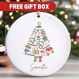 May include: White ceramic Christmas ornament with a green and red Christmas tree design. The tree is decorated with icons representing physical therapy, including balance, strength, mobility, and coordination. The text "PT" is in the center of the tree, and the name "Samantha" is at the bottom. The ornament is for sale with a free gift box.