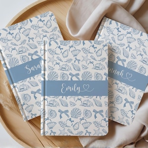 K&ouml;nnte beinhalten: Drei personalisierte Notizb&uuml;cher mit einem Muschel- und Schleifenmuster in Blau auf cremefarbenem Hintergrund. Jedes Notizbuch hat ein blaues Band mit einem Namen: Sarah, Emily und Hannah. Die Notizb&uuml;cher liegen auf einem Holztablett.