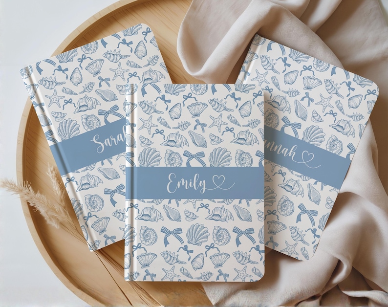 K&ouml;nnte beinhalten: Drei personalisierte Notizb&uuml;cher mit einem Muschel- und Schleifenmuster in Blau auf cremefarbenem Hintergrund. Jedes Notizbuch hat ein blaues Band mit einem Namen: Sarah, Emily und Hannah. Die Notizb&uuml;cher liegen auf einem Holztablett.