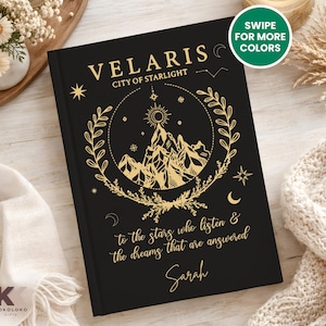 May include: Black journal with gold foil lettering and design. The cover reads "VELARIS CITY OF STARLIGHT" with a mountain and celestial design. The text "to the stars who listen & the dreams that are answered Sarah" is also on the cover. The journal has a matte finish.