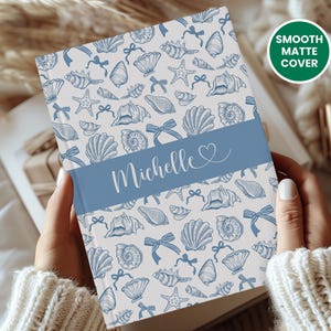 K&ouml;nnte beinhalten: Ein Notizbuch mit glatter, matter Oberfl&auml;che mit einem Muschelmuster in Blau und Wei&szlig;. Ein blaues Band in der Mitte zeigt den Namen "Michelle" in wei&szlig;er Schrift. Der Text "Smooth Matte Cover" befindet sich in einem gr&uuml;nen Kreis.