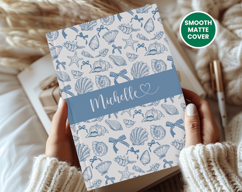 K&ouml;nnte beinhalten: Ein Notizbuch mit glatter, matter Oberfl&auml;che mit einem Muschelmuster in Blau und Wei&szlig;. Ein blaues Band in der Mitte zeigt den Namen "Michelle" in wei&szlig;er Schrift. Der Text "Smooth Matte Cover" befindet sich in einem gr&uuml;nen Kreis.