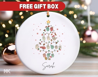 May include: White ceramic Christmas ornament with a green and red Christmas tree design. The tree is decorated with hand signs in a sign language alphabet. The ornament is personalized with the name "Sarah".