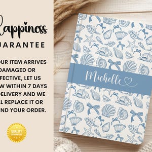 K&ouml;nnte beinhalten: Ein personalisiertes Notizbuch mit einem blau-wei&szlig;en Muschelmuster. Das Notizbuch hat ein blaues Band mit dem Namen "Michelle" in Schreibschrift. Der Text "Happiness Guarantee" ist ebenfalls sichtbar.