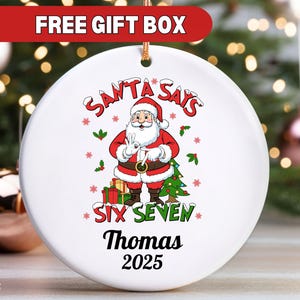 Könnte beinhalten: Runder, weißer Keramik-Weihnachtsschmuck mit einem Weihnachtsmann-Motiv. Der Schmuck trägt den Text "SANTA SAYS SIX SEVEN Thomas 2025". Er hat eine goldene Aufhängeschnur und den Text "FREE GIFT BOX" oben.