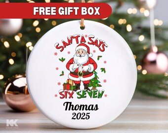 May include: White ceramic Christmas ornament with a Santa Claus graphic. The ornament has the text "SANTA SAYS SIX SEVEN Thomas 2025". The ornament is round and has a gold string for hanging. The words "FREE GIFT BOX" are at the top.